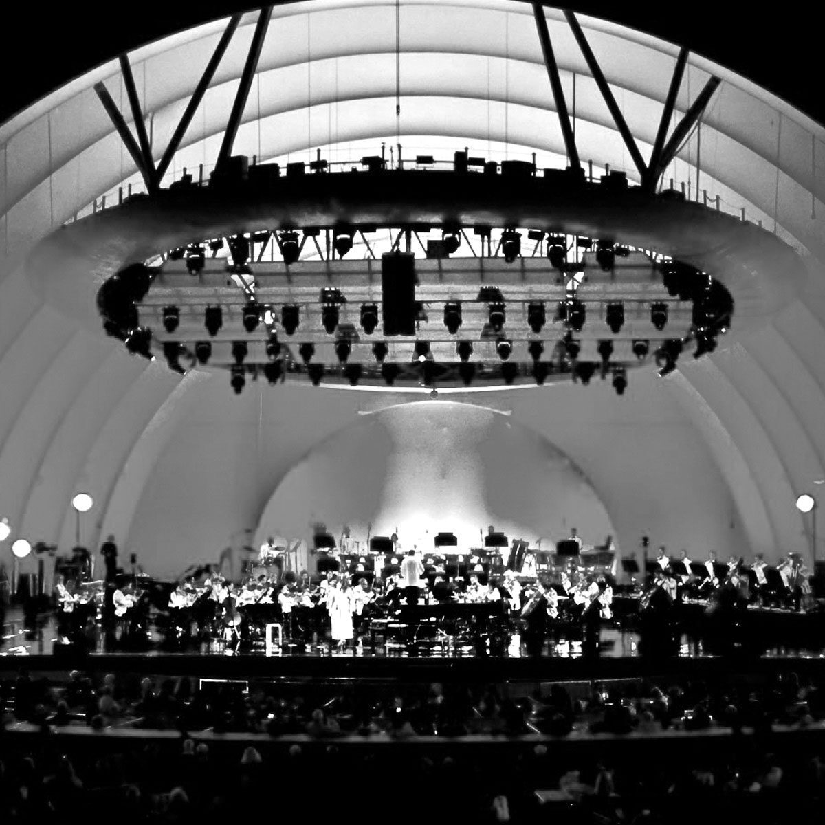 Orchestra playing on a large, arched stage with overhead lighting, creating a grand visual setting.